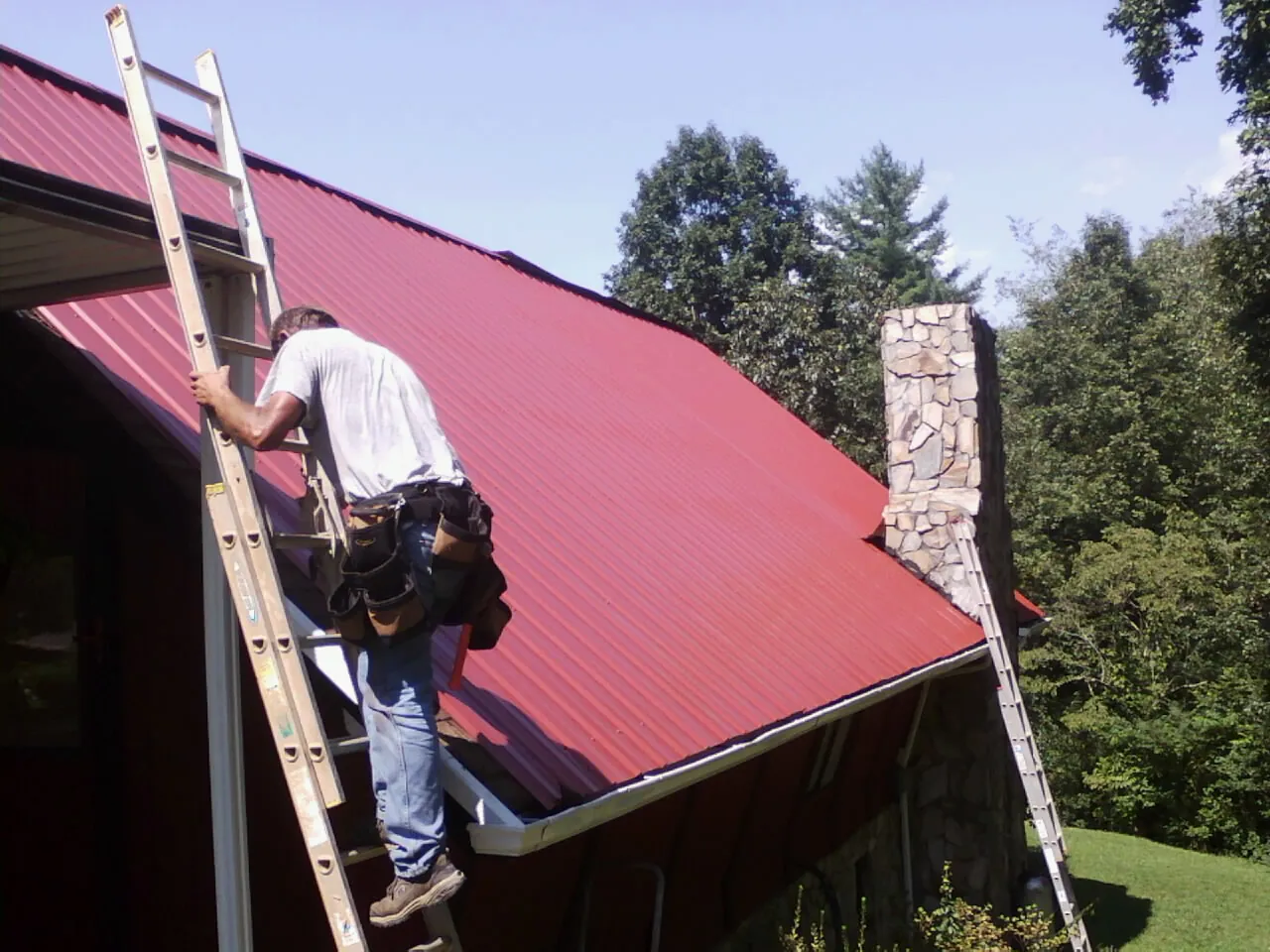 Roofer on a ladder beside a red metal roof and stone chimney — Soffit & Fascia Repair by Roofing Brothers in Black Eagle